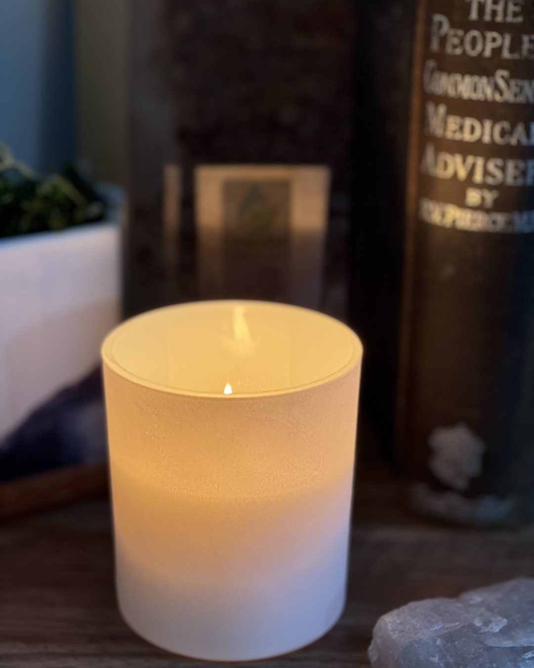 Lit candle in a white holder on a wooden surface with a blurred book in the background